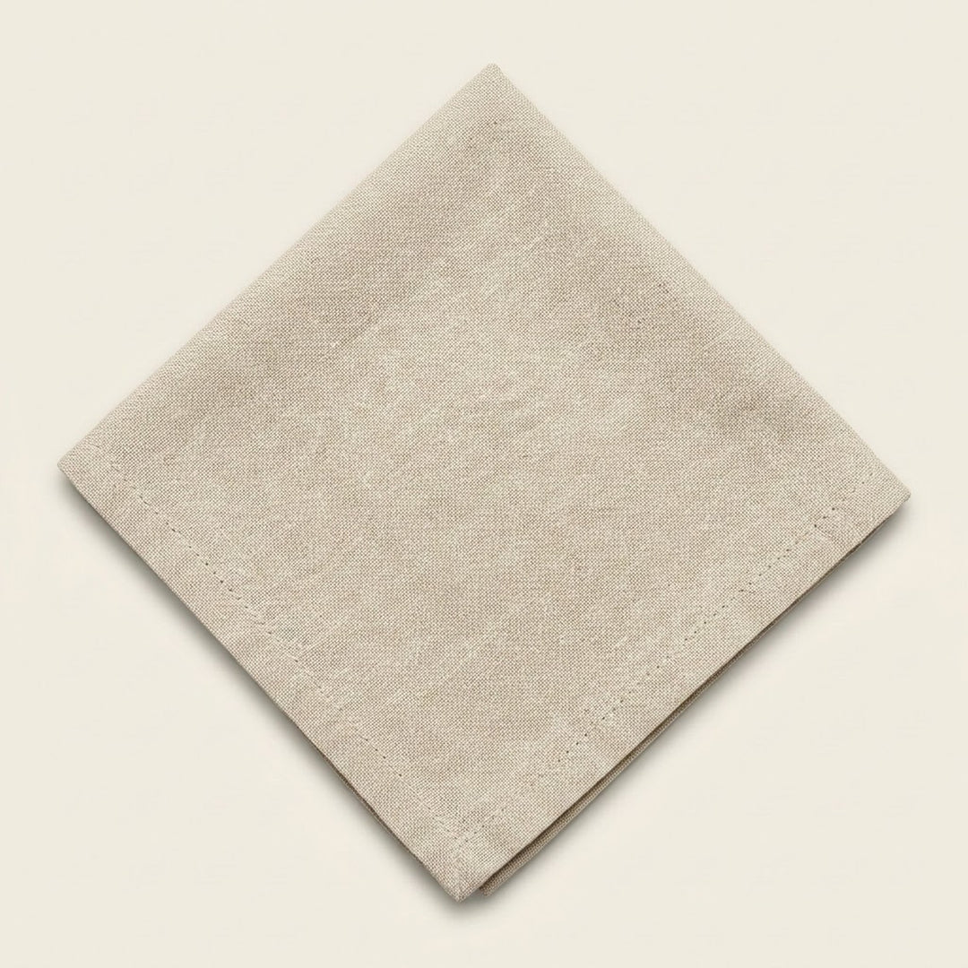 A folded beige linen napkin with a visible woven texture and stitched border, displayed on a solid cream background.