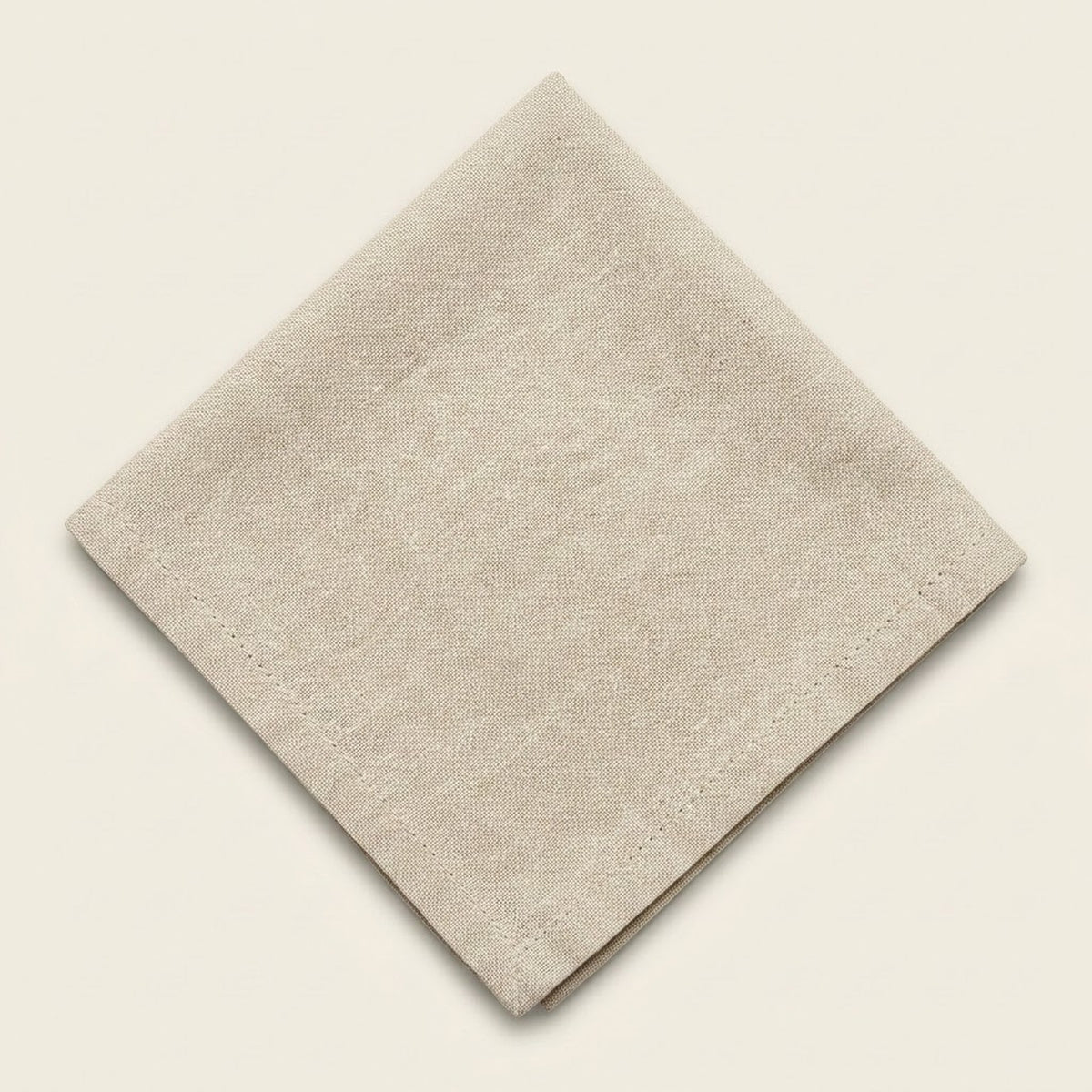A folded beige linen napkin with a visible woven texture and stitched border, displayed on a solid cream background.