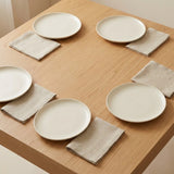Four round off-white ceramic plates arranged on a light wood table, each paired with a folded beige linen napkin.