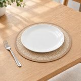 A white ceramic plate placed on a round woven beige placemat on a light wood table, with a silver fork to the left and a small plant in the upper left corner.
