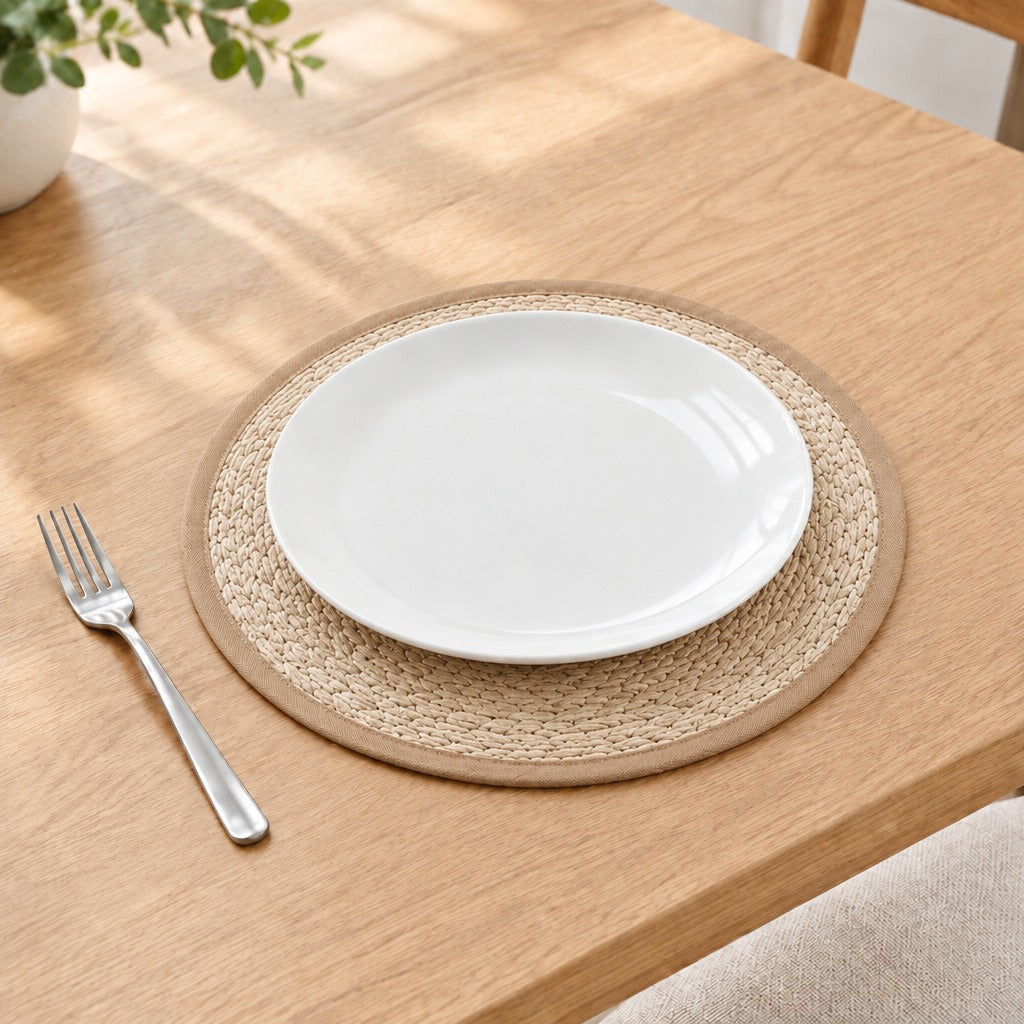 A white ceramic plate placed on a round woven beige placemat on a light wood table, with a silver fork to the left and a small plant in the upper left corner.