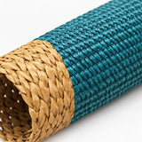 A close-up of a woven cylindrical mat or basket roll, showing a teal blue outer section and a natural tan straw inner section on a white background.