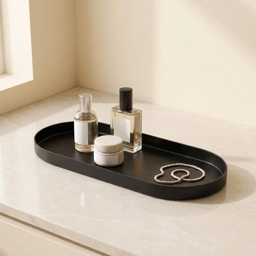 A black oval tray with a raised rim sits on a light marble countertop, holding a small bottle, a jar, and two heart-shaped rings. The scene is softly lit near a window with cream walls in the background.