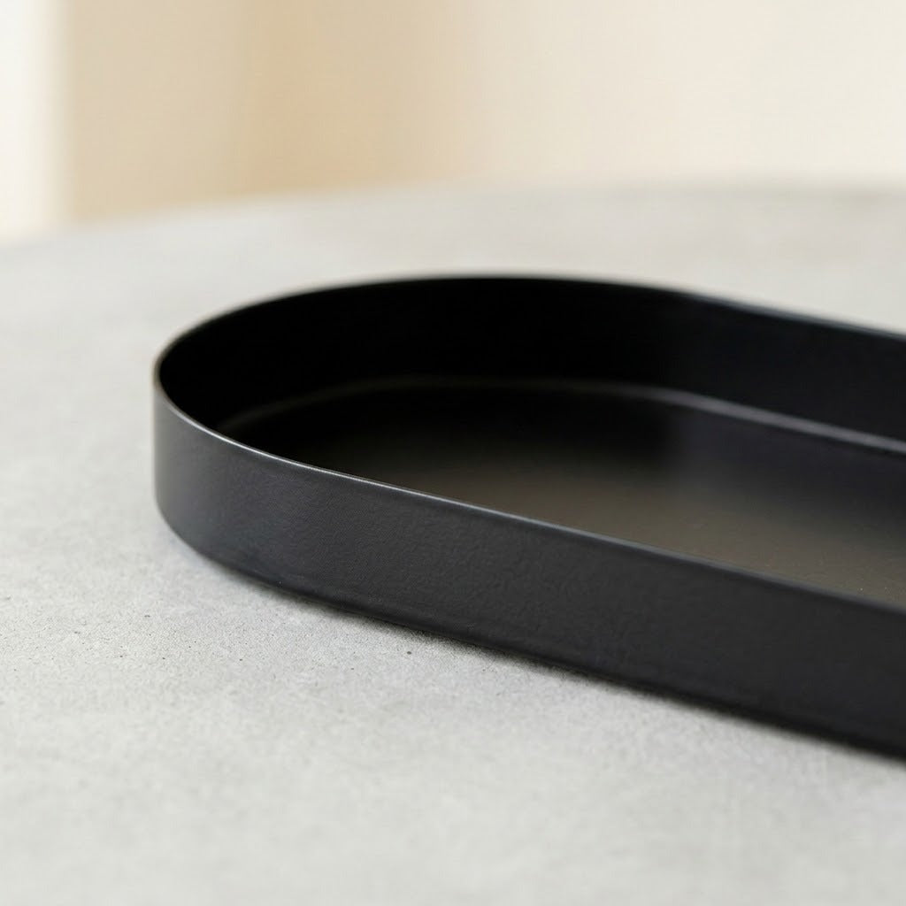 A close-up of a matte black metal tray with a rounded rectangular shape, resting on a light gray textured surface.