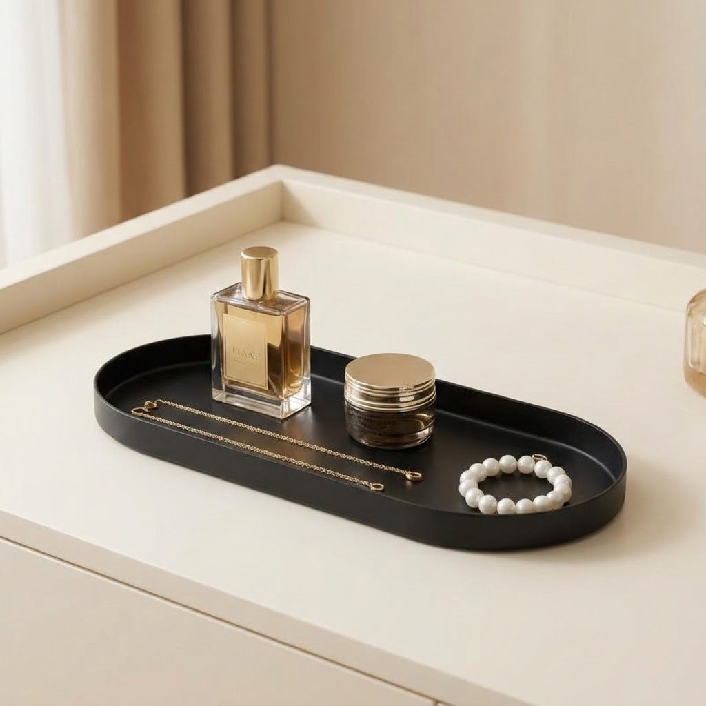 A black oval vanity tray with rounded corners sits on a cream-colored dresser. The tray holds a gold perfume bottle, a small gold jar, a gold chain necklace, and a white beaded bracelet.