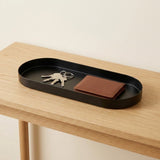 A black oval metal tray with rounded corners sits on a light wood table, holding a set of keys and a brown leather wallet.