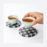 Two small tea cups filled with tea rest on round checkered coasters, with one cup being held by a hand. The coasters have a light blue and dark brown checkerboard pattern, shown on a clean white background.