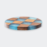 A round wooden board with a checkerboard pattern of natural brown wood and translucent blue resin squares, shown from a slight angle on a light gray background.