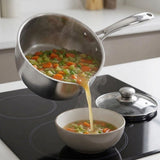 A stainless steel saucepan filled with vegetable soup is being poured into a white bowl on a black induction cooktop, with a glass lid resting nearby.