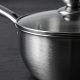 Close-up of a brushed stainless steel saucepan with a clear glass lid and metal handle, photographed on a dark textured surface.