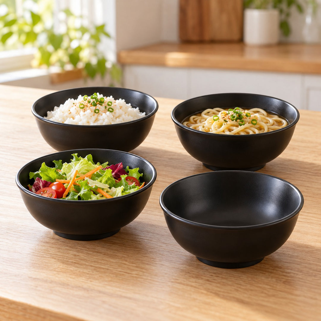 Four black Japanese-style bowls serve rice, noodles, salad and a side together on a kitchen table.