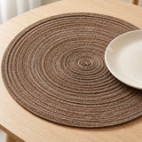 Brown round placemat shows its thick concentric braid beneath a simple ceramic plate on an oak table.