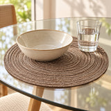 Brown woven polyester placemat cushions a serving bowl and glass on a bright glass-topped dining table.