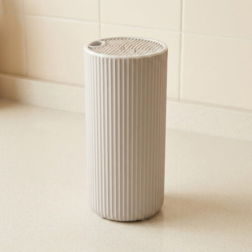 A ribbed cylindrical white container or dispenser standing on a light countertop against a tiled wall.
