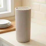 A ribbed cylindrical container or dispenser in a warm off-white/cream color, standing on a kitchen counter with plates and a tiled backsplash in the background.