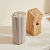A ribbed cylindrical white container with a slotted top sits beside a wooden block organizer on a light beige surface against a warm cream background.