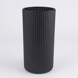 A black ribbed cylindrical container or vase with a fluted vertical texture, shown on a light gray background.