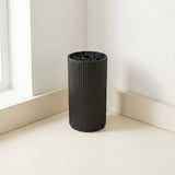 A black ribbed cylindrical container or vase standing on a cream-colored floor in the corner of a minimalist room with white walls.