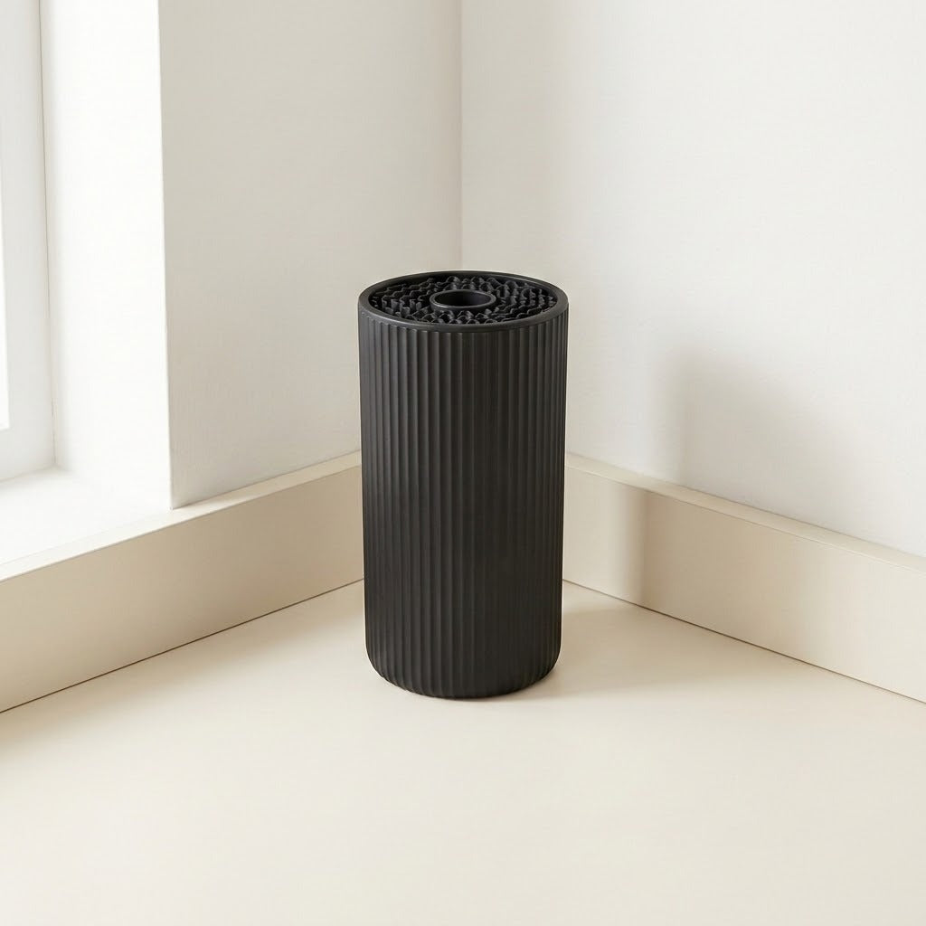 A black ribbed cylindrical container or vase standing on a cream-colored floor in the corner of a minimalist room with white walls.