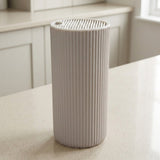 A ribbed cylindrical container or planter in a light warm gray or off-white color, standing on a speckled beige countertop with a blurred kitchen background.