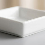 A close-up of a square white ceramic dish or shallow bowl with rounded corners, shown on a light gray surface with a softly blurred beige background.