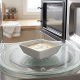 A white square bowl filled with a creamy yellow soup sits on a glass microwave turntable, with steam rising. The microwave door is open, and a blurred kitchen background is visible.