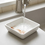 A square white ceramic dish with rounded corners sits on a kitchen counter under a running faucet, with water pooling inside.