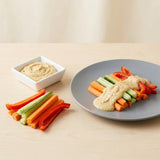 A plate of hummus dip with carrot sticks, cucumber sticks, and red bell pepper strips on a gray plate, alongside a small white bowl of hummus on a light wood table.