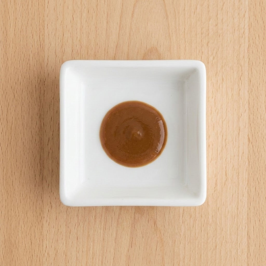A small white square ceramic dish containing a dollop of smooth brown sauce, centered on a light wood grain surface.