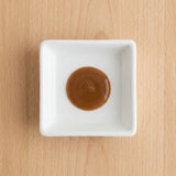 A small white square ceramic dish containing a dollop of smooth brown sauce, centered on a light wood grain surface.