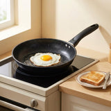 A black non-stick frying pan with a handle sits on a built-in stovetop in a kitchen, cooking a fried egg. A plate with toasted bread and a knife rests on the wooden countertop beside the stove.