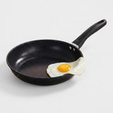 A black non-stick frying pan with a black handle, shown on a light gray background with a fried egg resting in the pan.
