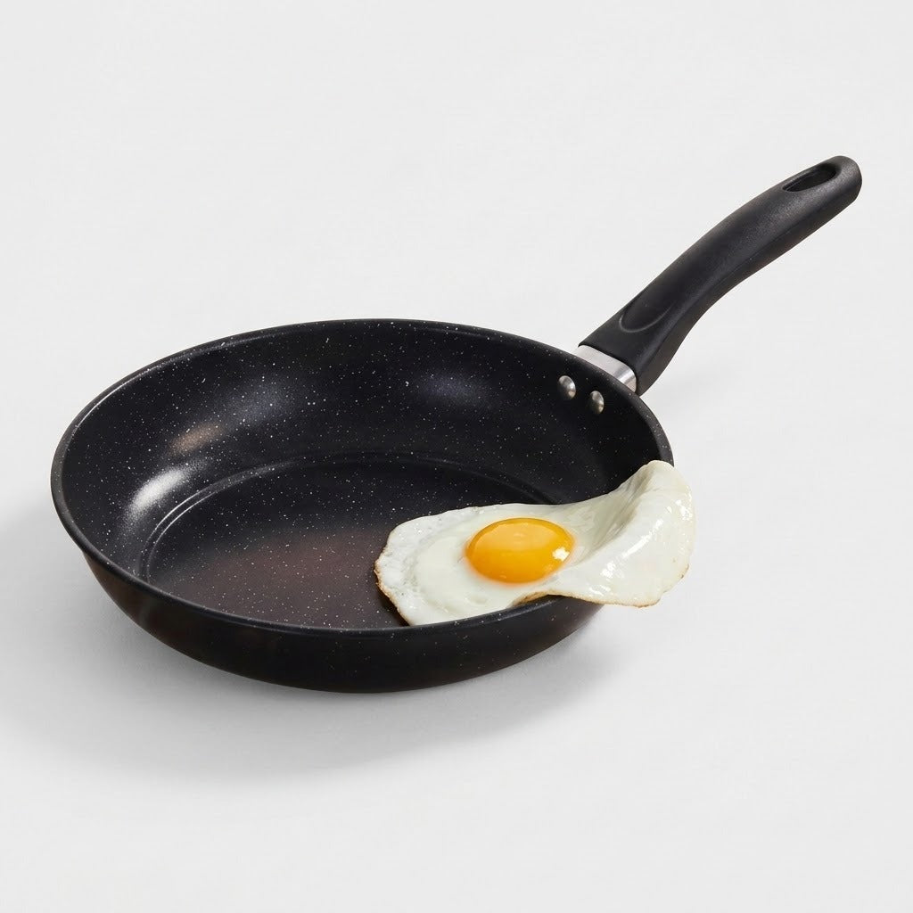 A black non-stick frying pan with a black handle, shown on a light gray background with a fried egg resting in the pan.