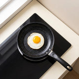 A black speckled non-stick frying pan with a handle sits on a black glass cooktop, cooking a sunny-side-up egg. The scene is shot from a high-angle perspective on a beige countertop near a window.