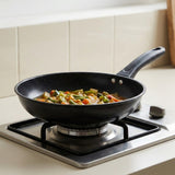 A black non-stick frying pan with a handle, shown on a gas stovetop with vegetables cooking inside. The background features a beige tiled wall and a light-colored countertop.