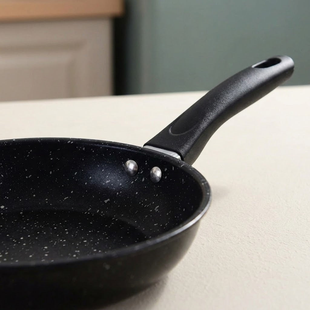 A close-up of a black non-stick frying pan with a speckled interior and a black handle, resting on a light-colored countertop in a kitchen setting.