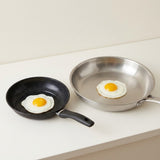 Two frying pans on a white countertop: a small black nonstick pan with a fried egg and a larger stainless steel pan with a fried egg, against a light beige wall.