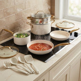A set of white ceramic cookware with gold-tone handles and lids is arranged on a kitchen counter beside a black induction cooktop. The cookware includes a double pot, a frying pan with tomato sauce, a smaller pan, and utensils, with steam rising from the pots in a kitchen setting.