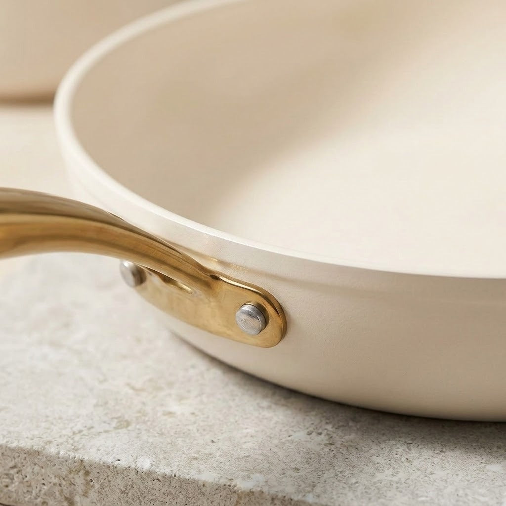 Close-up of a cream-colored nonstick frying pan with a gold-colored metal handle attachment, resting on a light gray stone surface.