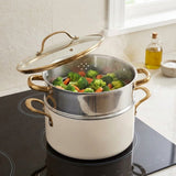 A cream-colored double pot with gold handles and a glass lid sits on a black induction cooktop in a kitchen setting, with vegetables steaming inside the top stainless steel pot.