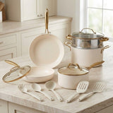 An 11-piece cream-colored cookware set with gold-tone handles and lids, arranged on a light marble kitchen countertop with matching utensils.
