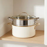 An off-white double boiler pot with a stainless steel inner pot, gold-toned handles and trim, and a glass lid, shown on a wooden shelf against a neutral background.