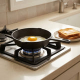 A black cast iron skillet with a frying egg on a lit gas stovetop in a kitchen, with toast on a plate nearby.