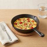 A black cast iron skillet filled with a tomato and herb omelet, set on a cork trivet on a light wood table with a folded cream napkin and fork, and a glass of water in the background.