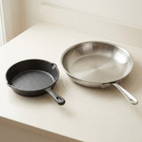 Two cookware pieces on a light cream countertop: a small black cast-iron skillet on the left and a larger stainless steel frying pan on the right.