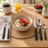 Black and silver flatware set arranged for breakfast beside a bowl on a light kitchen table.