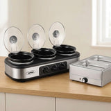 A three-compartment electric slow cooker with clear glass lids sits on a wooden countertop next to a stainless steel double compartment warmer, set against a kitchen background.