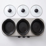 A stainless steel 3-burner electric hot plate with three black non-stick ceramic insert bowls, shown with three matching clear glass lids with black knobs on a white background.