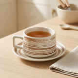 A white ceramic tea cup with a square handle and matching saucer, decorated with brown hand-painted horizontal brushstrokes. The cup is filled with tea and sits on a light wooden table with a folded beige napkin and a softly blurred background bowl and spoon.