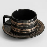 A black ceramic cup with a matching saucer, featuring horizontal distressed metallic gold and bronze stripes, set against a light gray background.